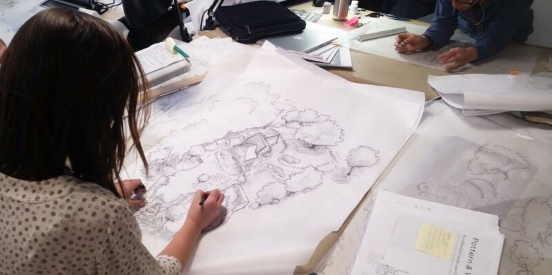 A view over a student's shoulder to the desk in front of her, where she is drawing a large design for a residential site.